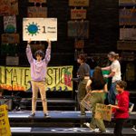 'Is My Microphone On?' by Jordan Tannahill, performed by Simon Langton Grammar School for Boys, Canterbury. An ensemble of young people surrounded by colourful placards and holding colourful placards that display slogans about the climate emergency.