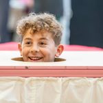 A young boy has his head through a hole in a trolley, he has curly hair on the top of his head that is blonde. He is smiling cheekily.