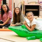 Three girls sit on the floor, left to right a young girl with black long curly hair wears black leggins and a pink t shirt, next to her a girl with long brown curly hair wears a green hoodie, next to her a girl with brown hair in French plaits wears a white tshirt, she is laying on an inflatable green crocodile.