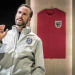 A man stands with his hand, aloft mid speech. He wears a grey England zip front, he has medium length beard and brown combed over hair. Behind him is a red England strip on a wooden door.