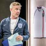 A man stands holding a clipboard, he wears a navy blue zipped top with the England emblem and the Nike tick. Underneath he is wearing a white tshirt. he has strawberry blonde hair, around his neck is a stop watch. Behind him is a white England strip shirt.