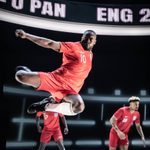 A man is mid air, mid high kick, his arm is outstretched his knees are in the air bent. He wears the red England strip. His face is focused. Behind him blurred are other players in the same strip.