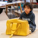 A young boy sits on the floor cross legged he has dark very curly hair and a wonderous expression. He sits in front of a large yellow handbag.