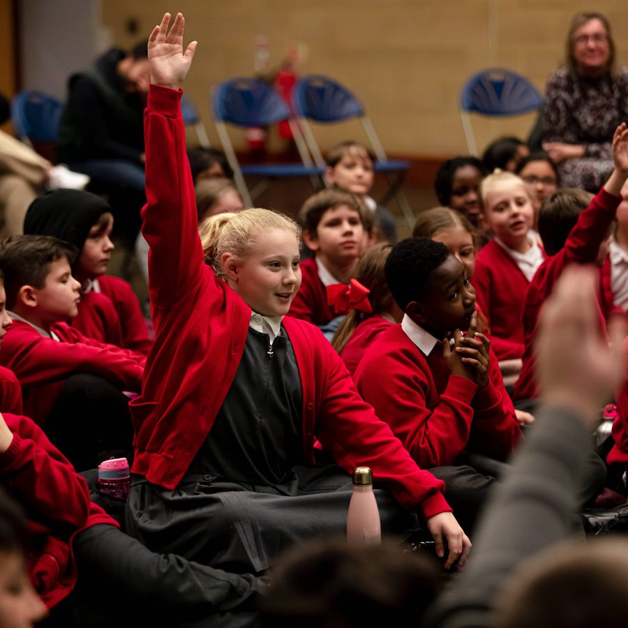 A group of primary school children, in the centre a young person with their hand in the air