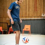 A young man wears over ear headphones, he is looking down at the ball he is playing keepy up with. He has afro hair which is shaved at the side but the top is long and peroxide dyed on the top. He wears a dark blue tshirt which says Utah State across the chest. He wears black jogger style shorts, he has white sport socks up to the ankle which have an orange band around them. He wears blue trainers.