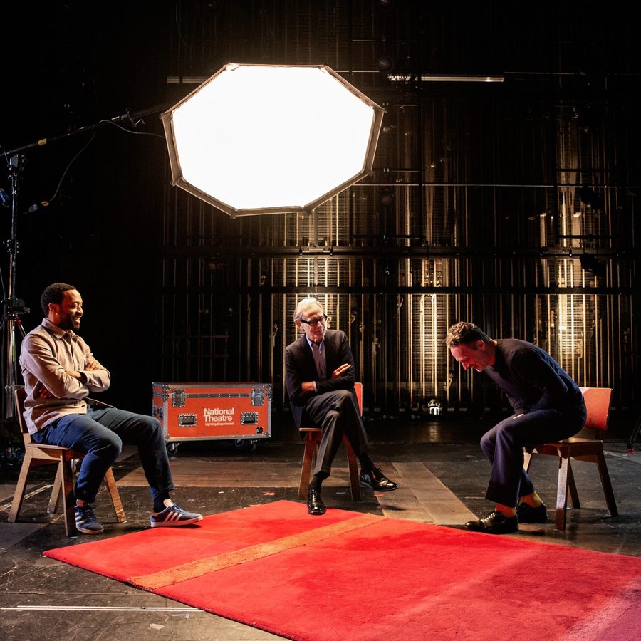 Three men sitting on chairs on a bare stage under a large whte light