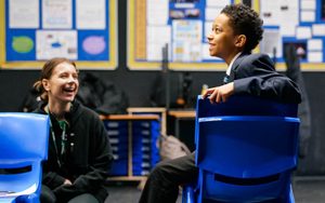 A young person sat on a plastic blue chair in a classroom, smiling with an adult member of staff smiling back