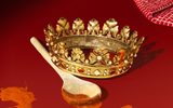 A gold and jewel encrusted crown rests on a wooden spoon that sits in a small pool of orange-coloured sauce, on a red surface. Sauce drips from part of the crown and a red and white checked cloth lies beyond the crown.
