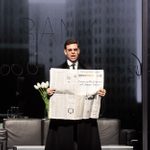 An actor in period attire stands, holiding an open newspaper, in front of a backdrop of skyscrapers. The furniture in the room is modern leather and steel.