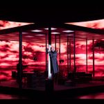 A man on a stage wearing a dark coat clings to a corner pillar of an open plan office. Behind are projections of strom clouds coloured red.
