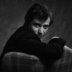 A black and white portrait photo of Johnny Flynn sitting in an arcmchair, turning round to look over the back, into the camera.