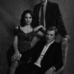 A black and white group portrait photo. Mark Gatiss stands behind an arcmchair in which sits Johnny Flynn, and on the arm of the chair sits a Tuppence Middleton.