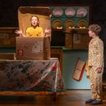 In a kitchen with a lot of packing boxes, a young girl wearing bright yellow bib-and-braces and long-sleeved top appears out of a large cardboard box on a table. A young boy in patterned pajamas stands to te side, lookig at her with his mouth sligtly ajar.